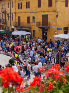 Una folla di persone riunita nella piazza principale di San Giorgio Canavese tra stand bianchi, palloncini e fiori rossi in primo piano.