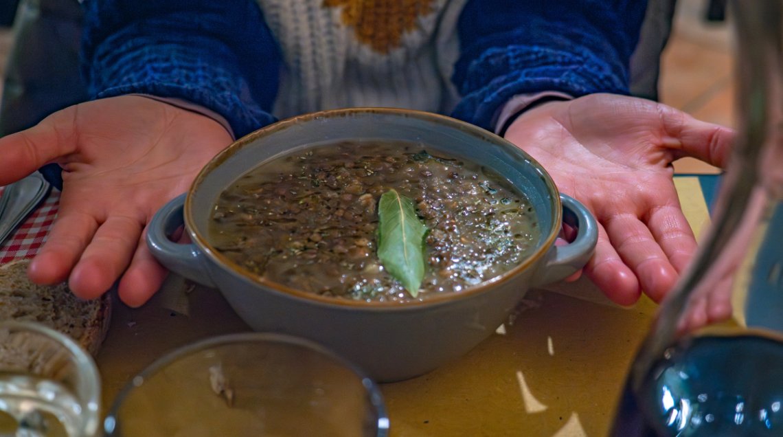Una ciotola di zuppa di lenticchie abruzzese guarnita con una foglia di alloro, presentata tra le mani aperte di una persona.