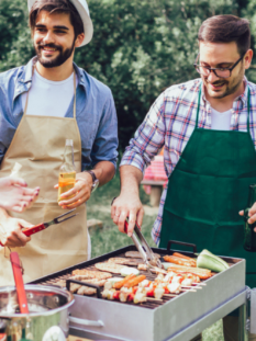 Un gruppo di amici fanno il barbecue all'aperto, due amici curano il grill e gli altri scherzano