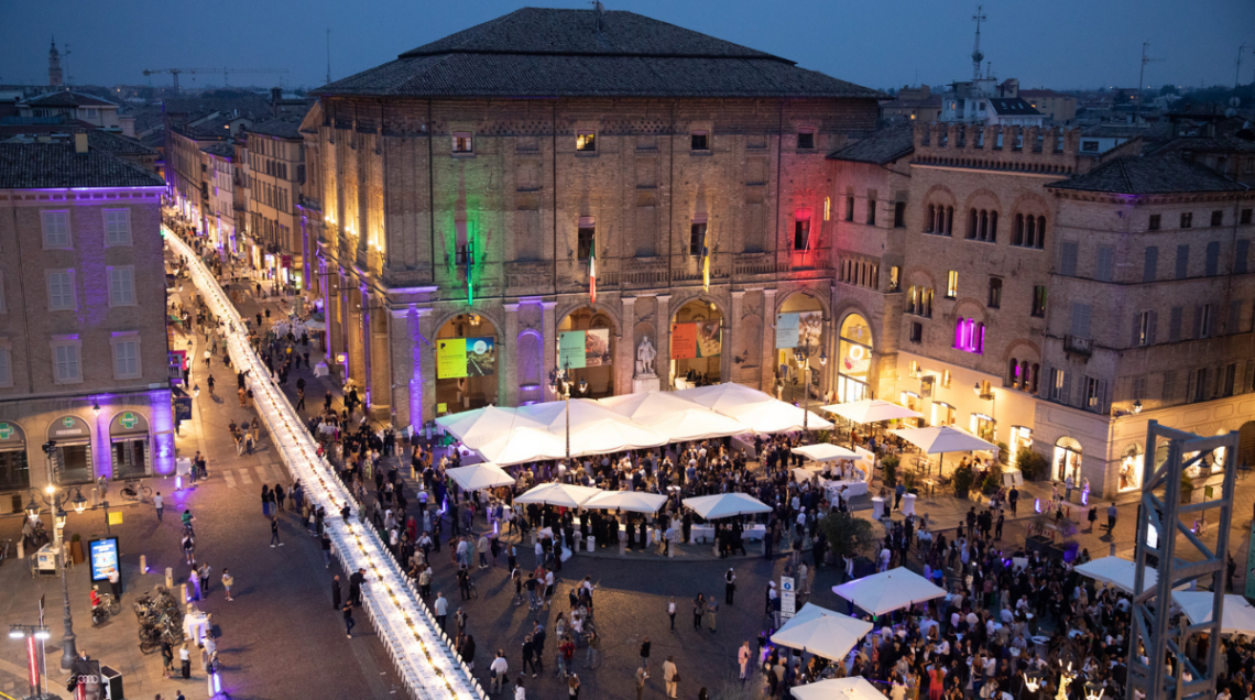 Veduta dall'alto di Piazza Garibaldi a Parma durante la Cena dei Mille, con i tavoli bianchi allestiti, ombrelloni, e il Palazzo del Governatore illuminato con il tricolore italiano.