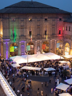 Veduta dall'alto di Piazza Garibaldi a Parma durante la Cena dei Mille, con i tavoli bianchi allestiti, ombrelloni, e il Palazzo del Governatore illuminato con il tricolore italiano.