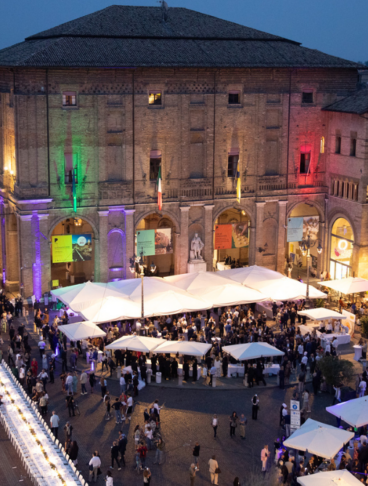 Veduta dall'alto di Piazza Garibaldi a Parma durante la Cena dei Mille, con i tavoli bianchi allestiti, ombrelloni, e il Palazzo del Governatore illuminato con il tricolore italiano.