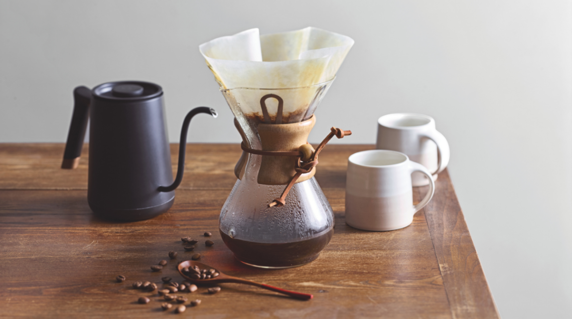 Caraffa Chemex in vetro con collare in legno su un tavolo rustico accanto a un bollitore moderno nero e chicchi di caffè.