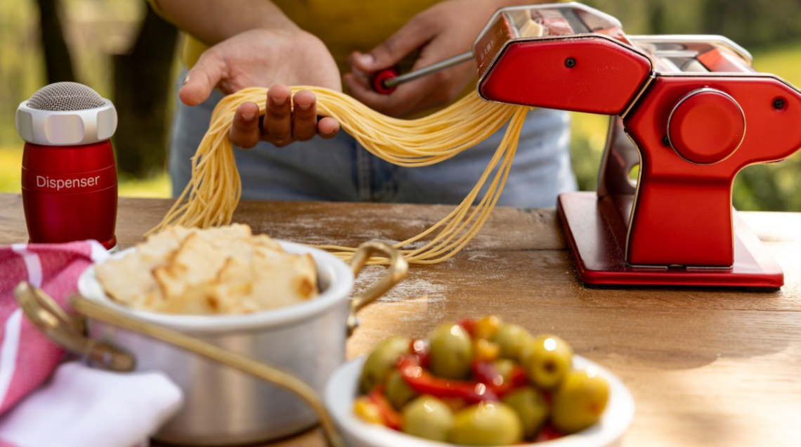 Macchina tirapasta Marcato Atlas+ di colore rosso metallizzato che realizza spaghetti freschi all'aperto mentre una persona raccoglie la pasta sul palmo della mano.