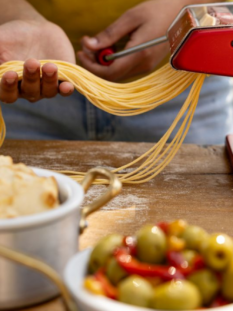 Macchina tirapasta Marcato Atlas+ di colore rosso metallizzato che realizza spaghetti freschi all'aperto mentre una persona raccoglie la pasta sul palmo della mano.