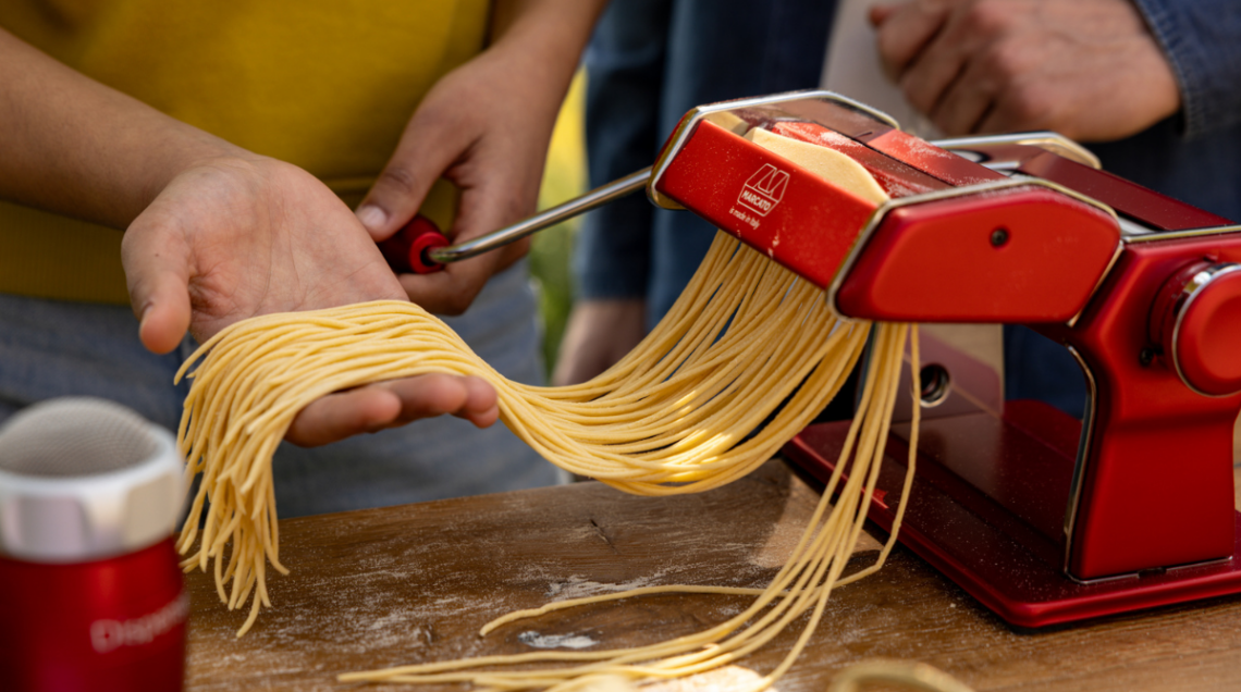 Macchina tirapasta Marcato Atlas+ di colore rosso metallizzato che realizza spaghetti freschi all'aperto mentre una persona raccoglie la pasta sul palmo della mano.