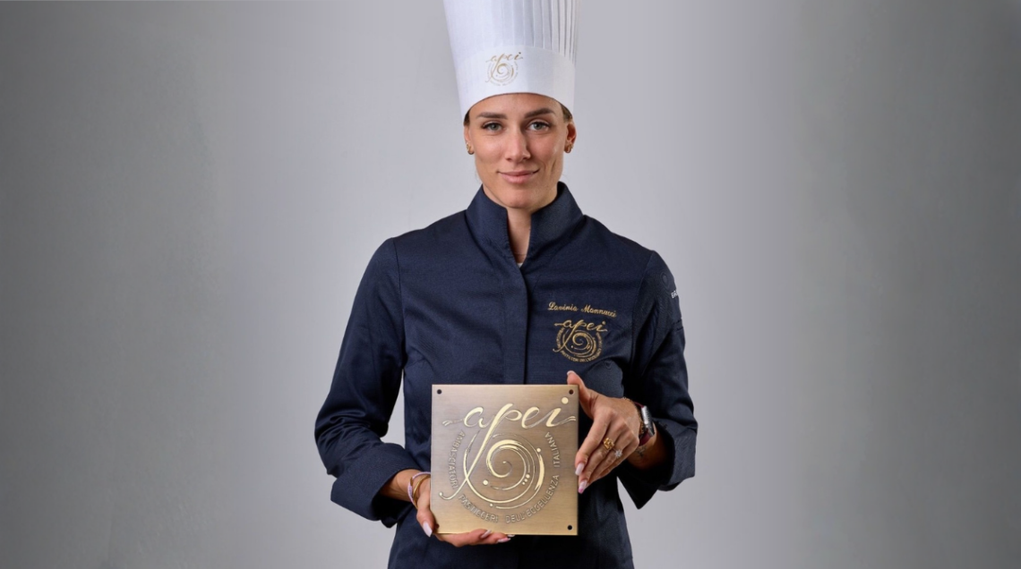 Lavinia Mannucci in divisa scura da chef accademico mostra la targa ufficiale dorata dell'associazione APEI (Ambasciatori Pasticceri dell'Eccellenza Italiana).