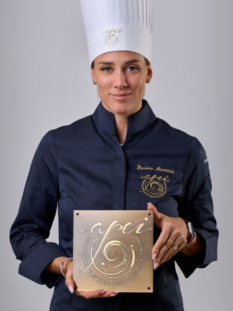 Lavinia Mannucci in divisa scura da chef accademico mostra la targa ufficiale dorata dell'associazione APEI (Ambasciatori Pasticceri dell'Eccellenza Italiana).