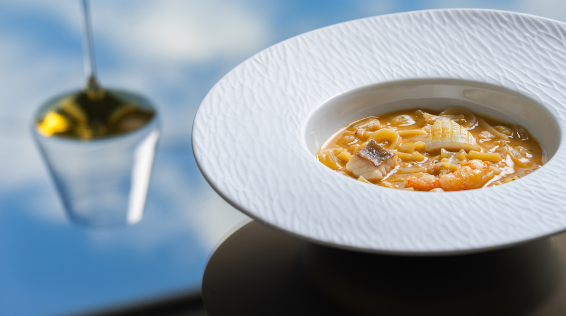 Un piatto fondo bianco con una zuppa di mare, pasta mista, crostacei e pesci di scoglio, adagiato su una superficie riflettente azzurra