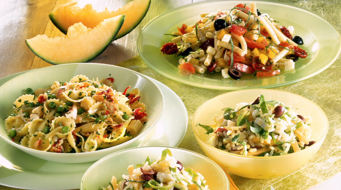 piatti e ciotole di insalate fredde di riso e cereali disposte su una superficie piana, con due fette di melone a lato