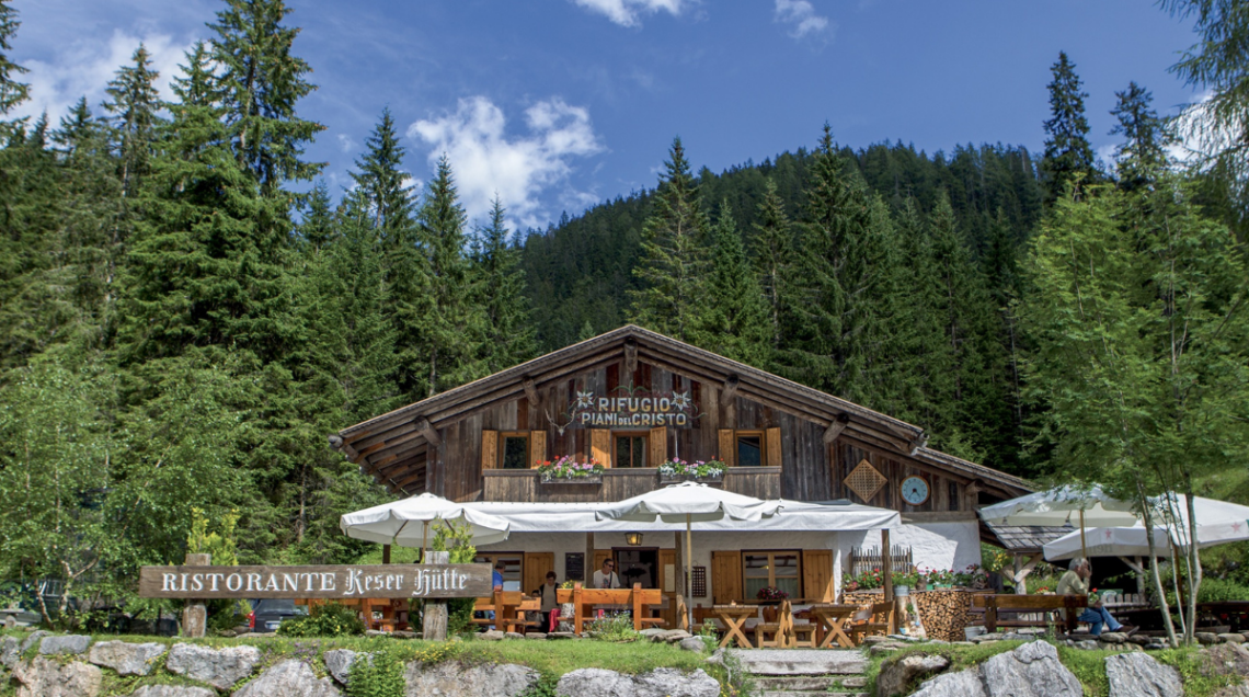 Facciata in legno del Rifugio Piani del Cristo con tavoli all'aperto e ombrelloni bianchi ai piedi di una fitta foresta di pini. 