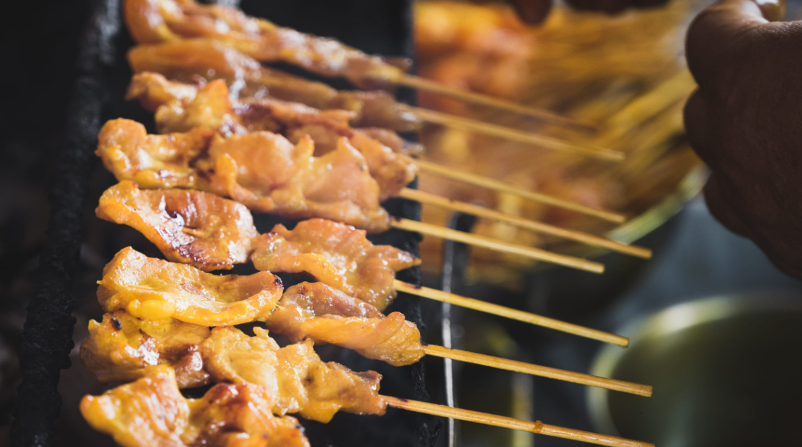 Spiedini di legno con pezzetti dorati di carne di maiale, sono cotti distesi sull'apposito grill satay, uno vicino all'altro