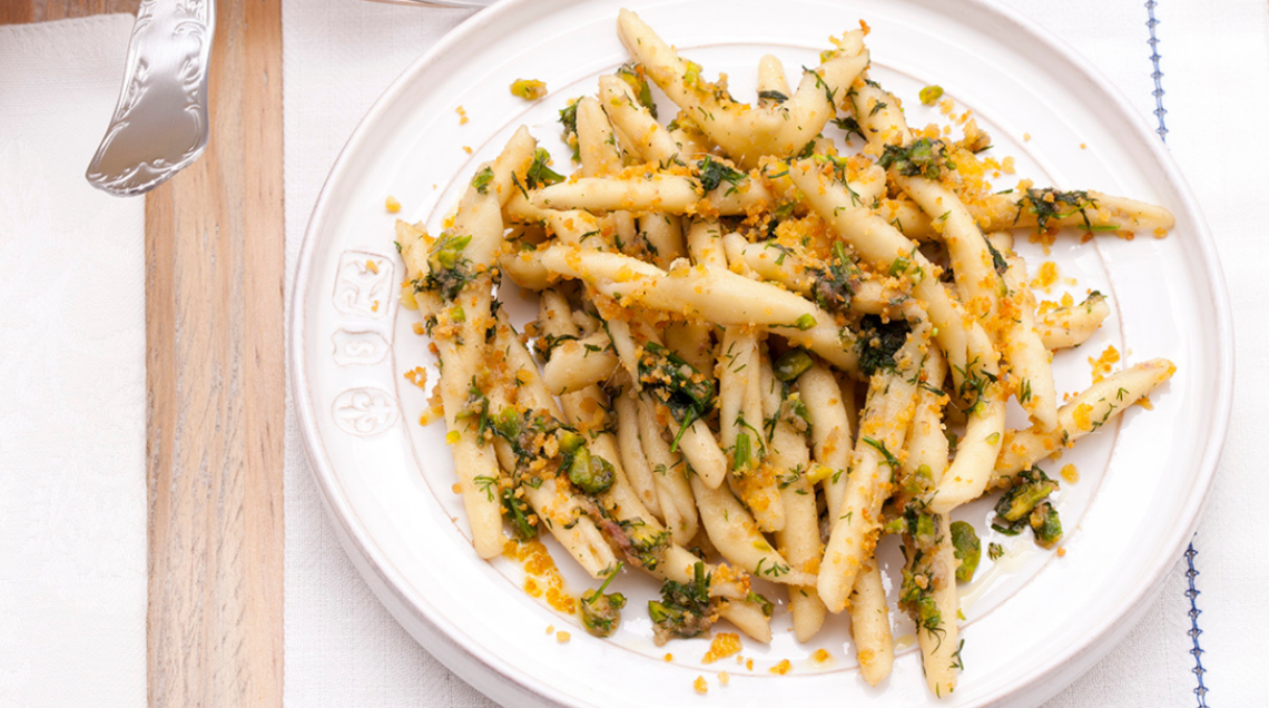 Un piatto bianco di ceramica con busiate condite con pesto, granella di mandorle o pistacchi ed erbe aromatiche.
