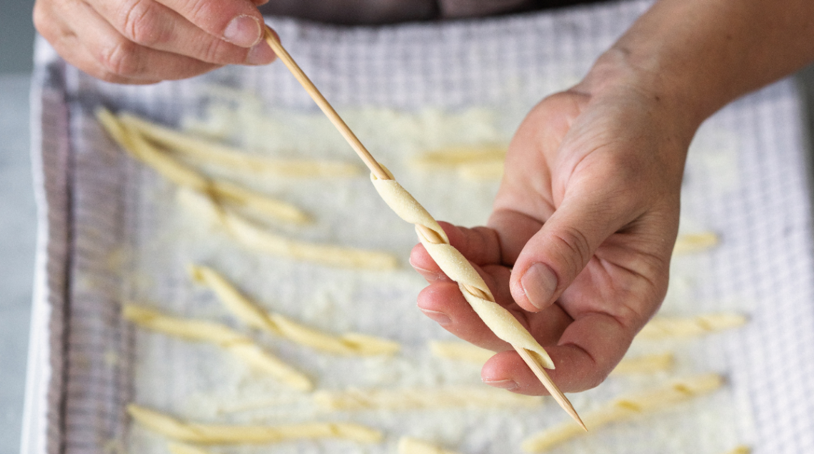Mani che arrotolano la pasta fresca siciliana attorno a un bastoncino di legno per fare le busiate artigianali.