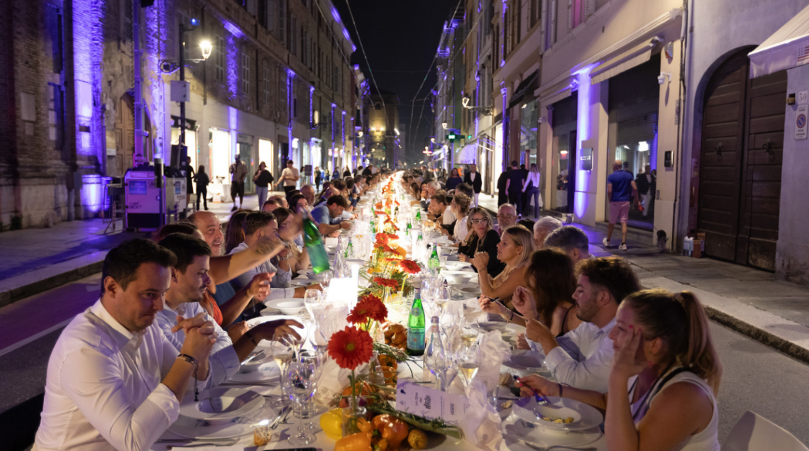 Ospiti seduti lungo l'infinita tavolata della Cena dei Mille a Parma di notte, decorata con fiori rossi e candele, tra i palazzi storici illuminati di viola.