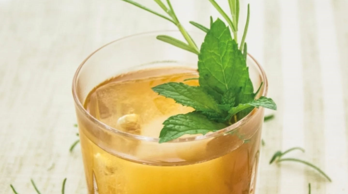 Un bicchiere basso di vetro lavorato contenente un cocktail dal colore ambrato con un grande cubetto di ghiaccio, guarnito elegantemente con un rametto di rosmarino fresco e foglie di menta, appoggiato su una tovaglietta a righe.