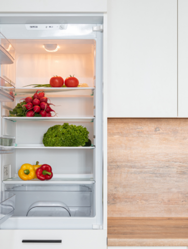 Frigorifero moderno da incasso aperto e illuminato, con verdure fresche disposte sui vari ripiani di vetro in una cucina bianca con paraschizzi in legno