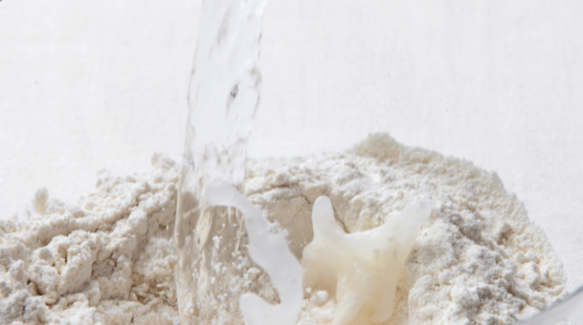 flusso di acqua che cade nel centro di un cumulo di farina bianca in una ciotola di vetro trasparente. Il movimento crea schizzi bianchi.