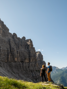 Coppia di escursionisti con zaini ammira un paesaggio di alte montagne rocciose e guglie calcaree sotto un cielo azzurro e soleggiato.