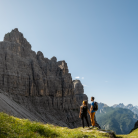 Coppia di escursionisti con zaini ammira un paesaggio di alte montagne rocciose e guglie calcaree sotto un cielo azzurro e soleggiato.