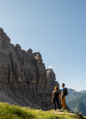 Coppia di escursionisti con zaini ammira un paesaggio di alte montagne rocciose e guglie calcaree sotto un cielo azzurro e soleggiato.