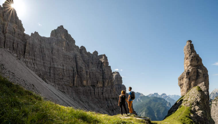 Coppia di escursionisti con zaini ammira un paesaggio di alte montagne rocciose e guglie calcaree sotto un cielo azzurro e soleggiato.