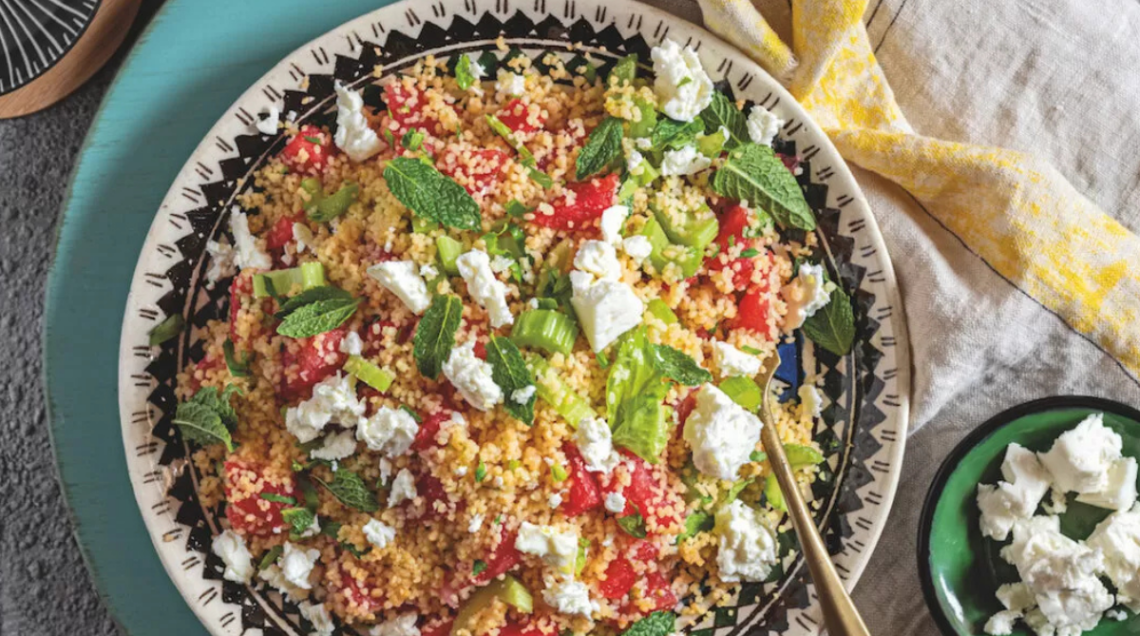 insalata di cous cous con anguria, sedano e feta in un grande piatto disposto su un tavolo con dentro un cucchiaio, con a lato una piccola ciotola di feta