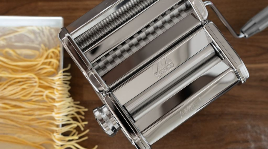 Macchina sfogliatrice manuale Marcato Atlas+ in acciaio cromato lucido posizionata sul piano di una cucina moderna con un vassoio di pasta fresca accanto.