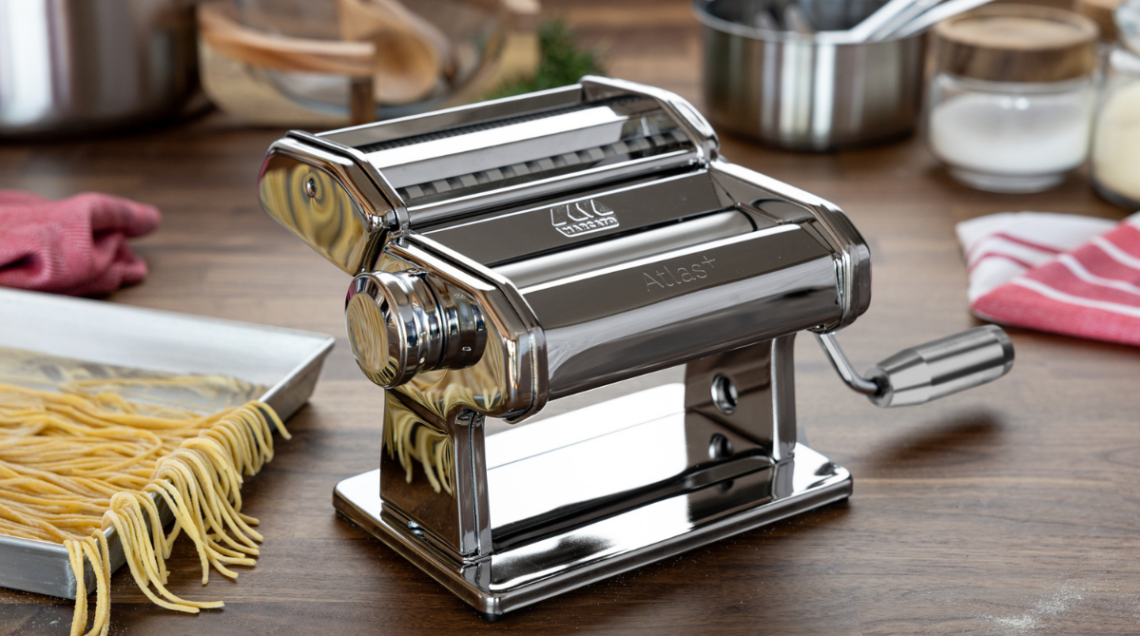 Macchina sfogliatrice manuale Marcato Atlas+ in acciaio cromato lucido posizionata sul piano di una cucina moderna con un vassoio di pasta fresca accanto.