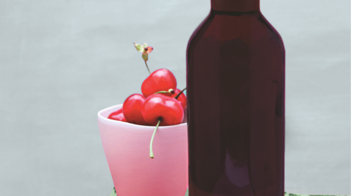 Una bottiglia di liquore scuro accanto a un bicchiere colmo di ciliegie fresche su una tovaglia verde a motivi vegetali.