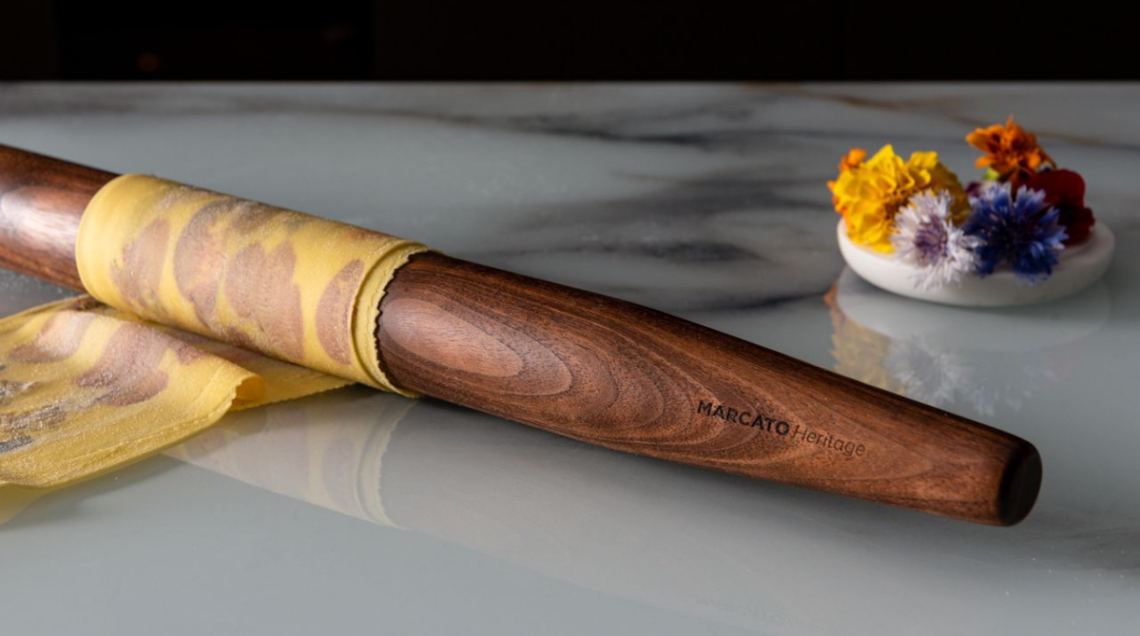 Mattarello francese Marcato in legno massello scuro che arrotola una sfoglia di pasta liscia su un top da cucina in marmo bianco.