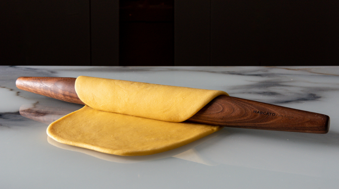 Mattarello francese Marcato in legno massello scuro che arrotola una sfoglia di pasta liscia su un top da cucina in marmo bianco.