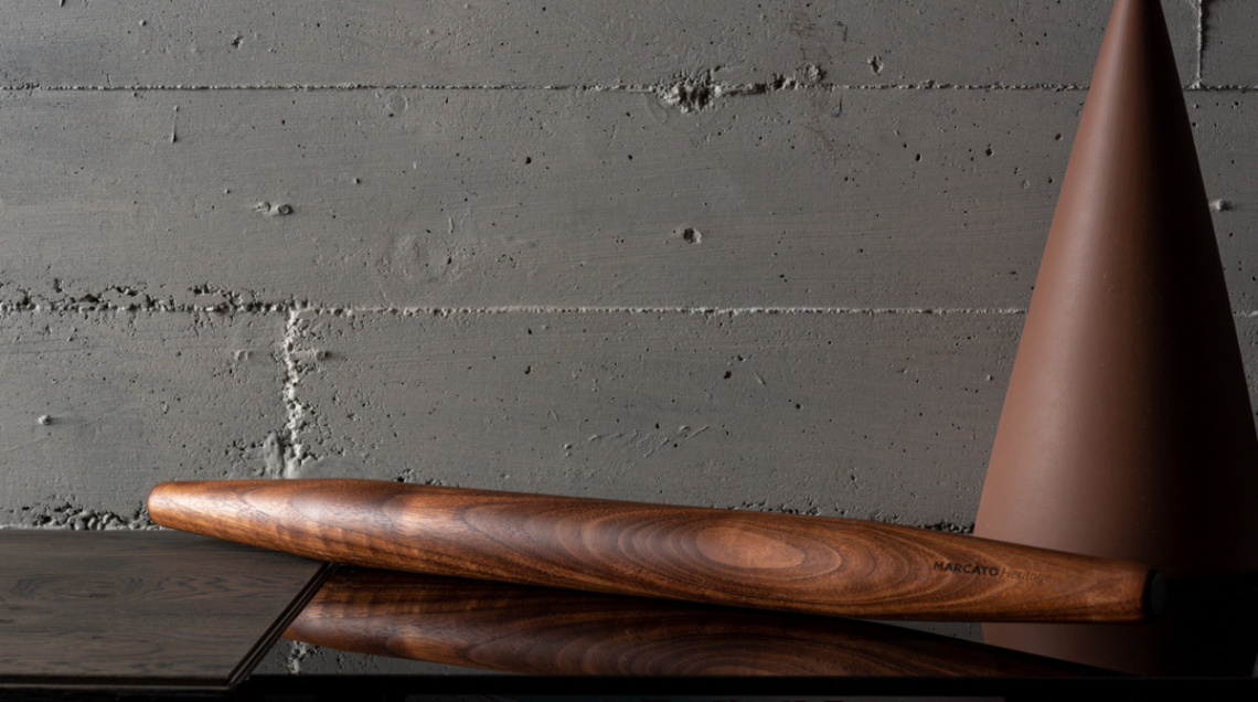 Il Mattarello Marcato della linea Heritage in legno massello scuro adagiato su un piano riflettente nero davanti a una parete grigia in cemento.