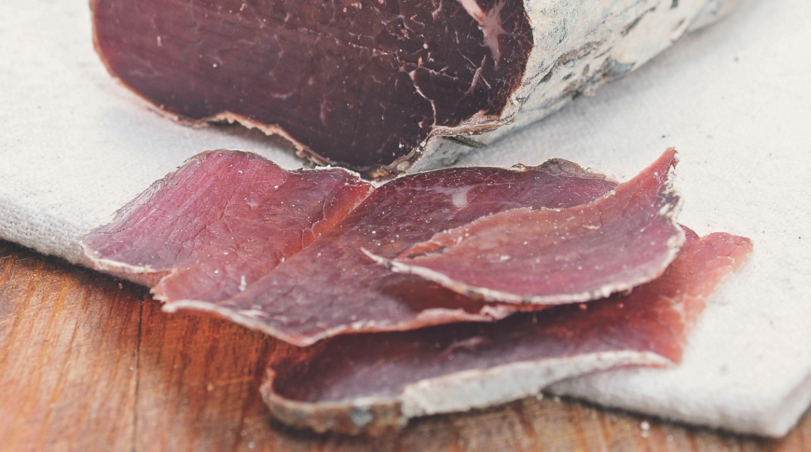 Fette sottili di mocetta valdostana disposte su carta alimentare accanto al pezzo intero di carne essiccata.