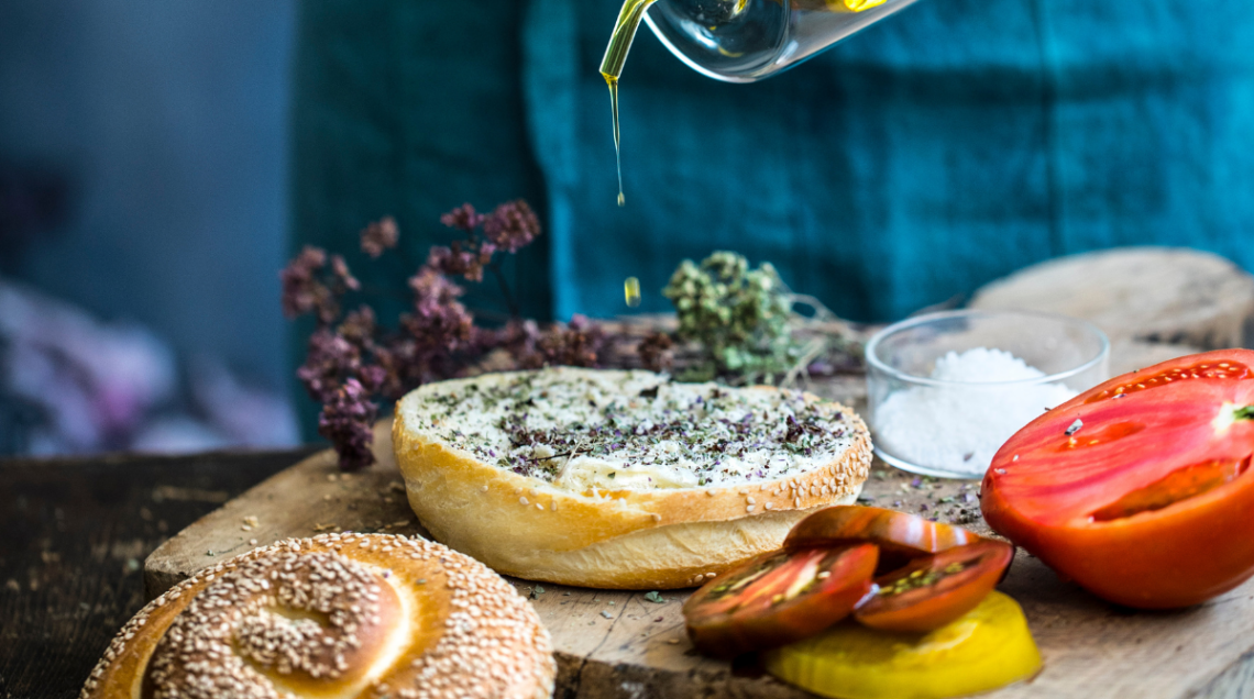 Una vista a media inquadratura che mostra la preparazione del pani cunzatu. Una persona in grembiule versa olio d'oliva da un beccuccio di vetro su un panino al sesamo precedentemente cosparso di origano secco. Sul tagliere di legno ci sono pomodori affettati, sale marino e origano secco.