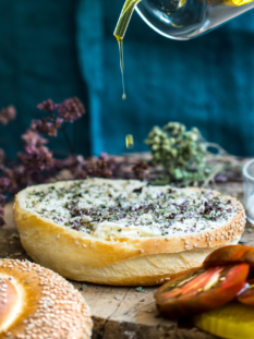 Una vista a media inquadratura che mostra la preparazione del pani cunzatu. Una persona in grembiule versa olio d'oliva da un beccuccio di vetro su un panino al sesamo precedentemente cosparso di origano secco. Sul tagliere di legno ci sono pomodori affettati, sale marino e origano secco.