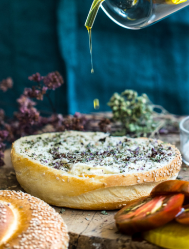 Una vista a media inquadratura che mostra la preparazione del pani cunzatu. Una persona in grembiule versa olio d'oliva da un beccuccio di vetro su un panino al sesamo precedentemente cosparso di origano secco. Sul tagliere di legno ci sono pomodori affettati, sale marino e origano secco.