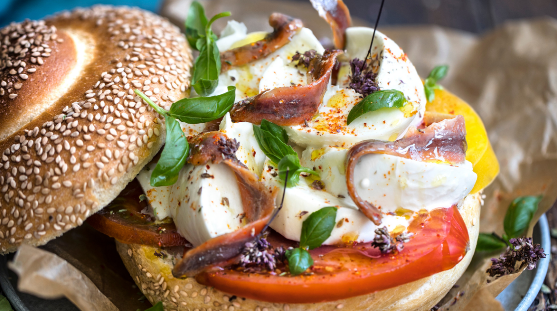 Un primo piano ravvicinato e rustico di un pani cunzatu siciliano completo, farcito con fette di pomodoro rosso, fette di mozzarella fresca, acciughe salate arrotolate, foglie e fiori di basilico fresco e una spolverata di peperoncino, servito su carta da forno.