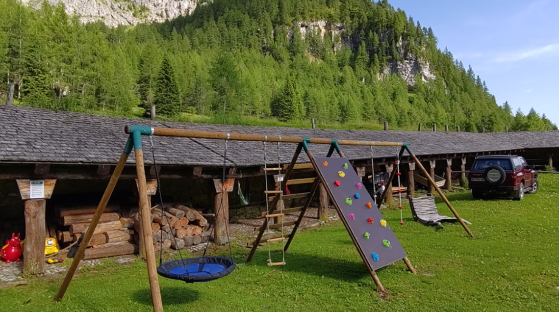 Area giochi in legno con altalena e parete da arrampicata davanti a una struttura alpina tradizionale immersa nel verde delle montagne friulane.