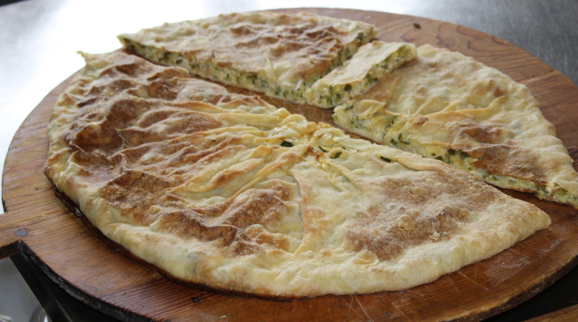 Una grande torta salata rotonda tradizionale ligure (torta verde) con sfoglia dorata e croccante, tagliata a fette e presentata su un grande tagliere circolare di legno scuro. 