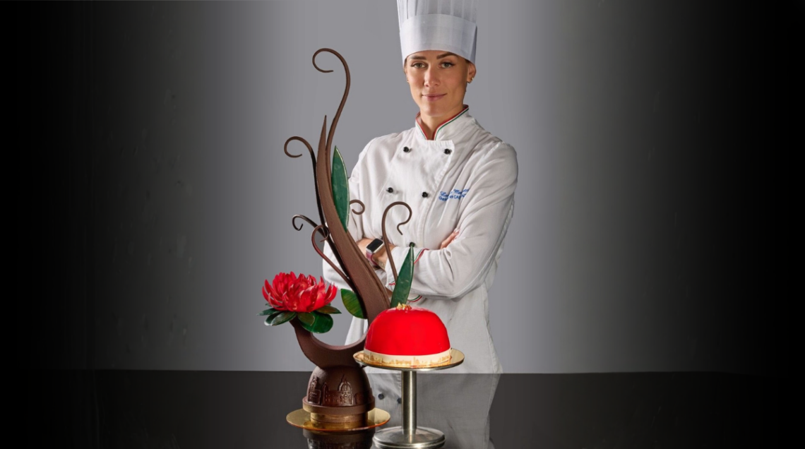 La chef Lavinia Mannucci in divisa bianca dietro alla sua opera: lo zuccotto fiorentino rosso cupola e una scultura artistica in cioccolato.