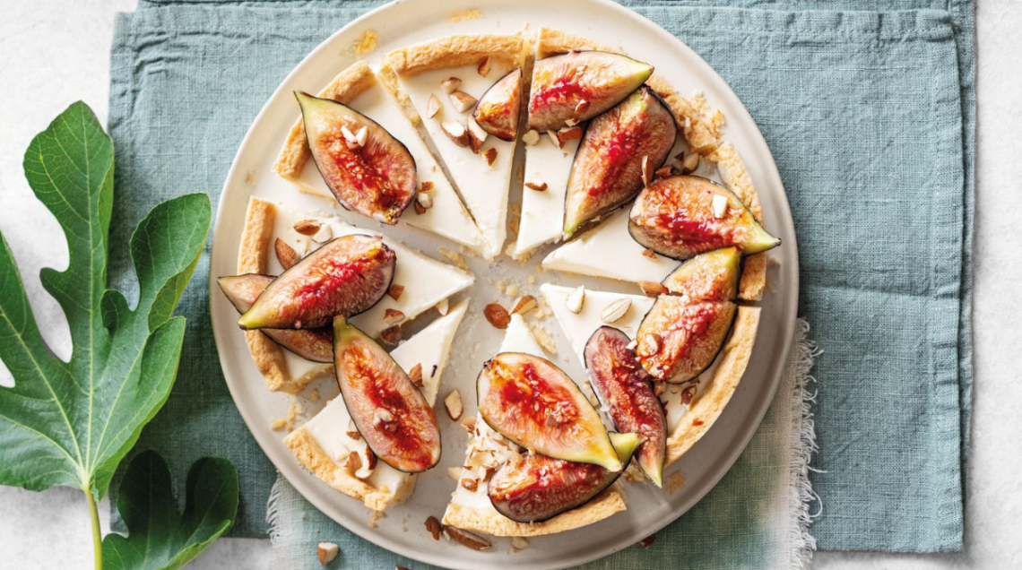 crostata con panna cotta, fichi e mandorle tagliata a fette, su piatto bianco e sopra un canovaccio azzurro. Alla sua sinistra una foglia di fico