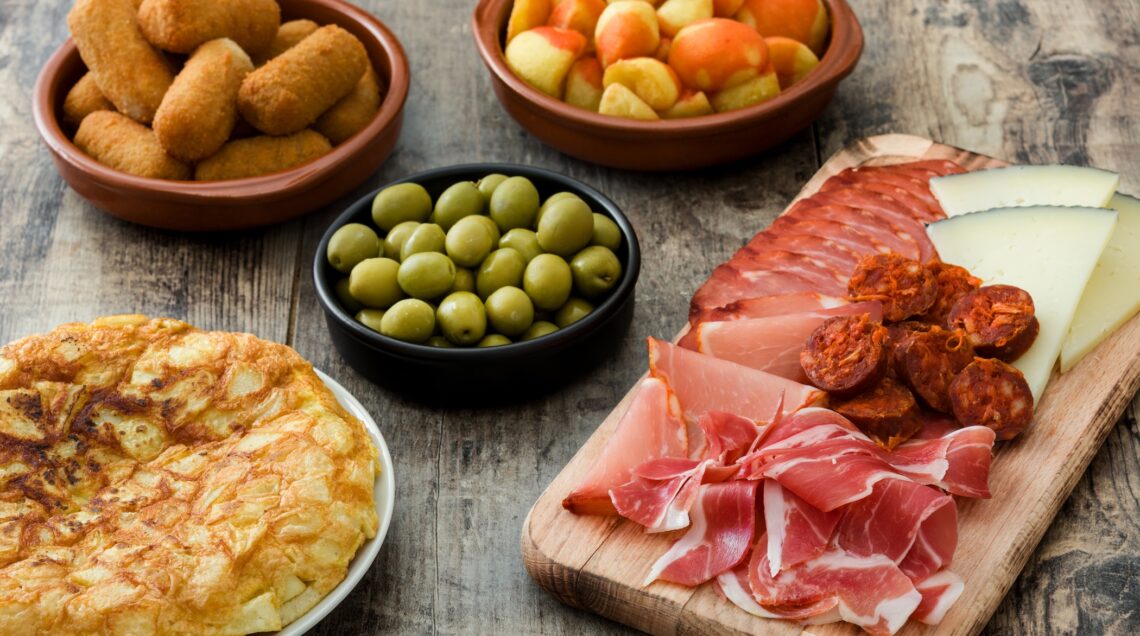 Un tavolo con tutte le Tapas tradizionali andaluse: ci sono ciotole con crocchette di patate, olive, tortilla, patatas bravas e un tagliere con affettati, jamon e formaggi