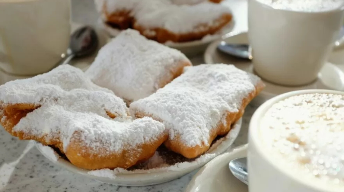 Beignets spolverati di zucchero a velo su piattino bianco, affianco a due cappuccini