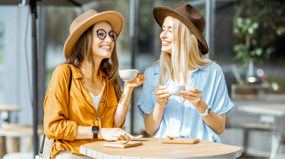 Due ragazze sorridenti, una mora e una bionda, degustano due caff&egrave; appoggiate a un tavolo di legno chiaro