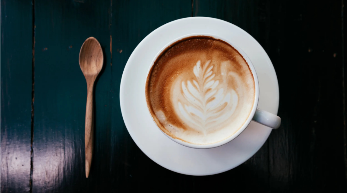 Cappuccino in tazza bianca su sfondo scuro. Alla sua sinistra un cucchiaino di legno
