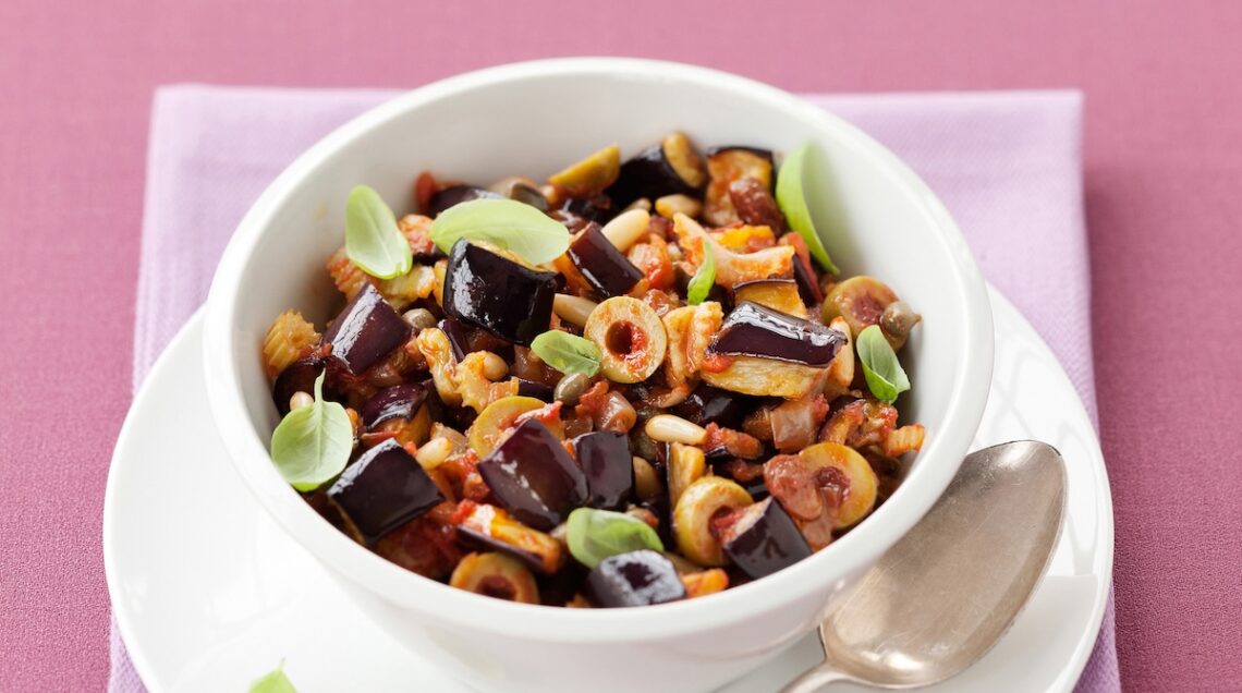 Caponata di melanzane all'interno di una ciotola bianca, sopra un piatto piano bianco con cucchiaio di metallo. Sotto un tovagliolo rosa