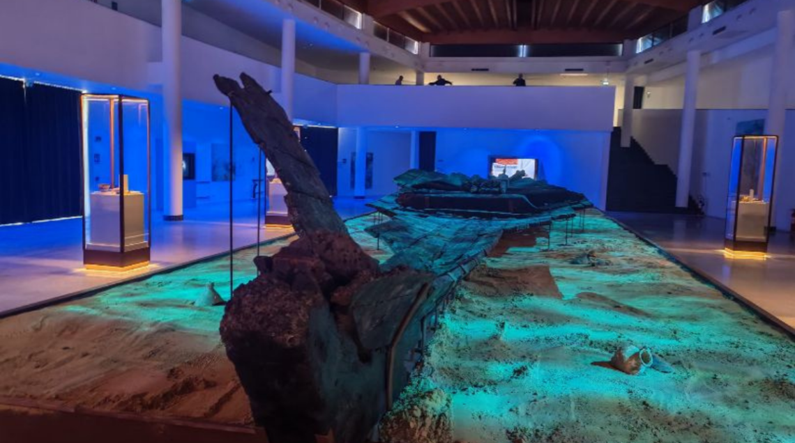 Interno del Museo dei Relitti Greci a Gela con lo scafo di un'antica nave adagiato sulla sabbia sotto luci azzurre e teche espositive. 