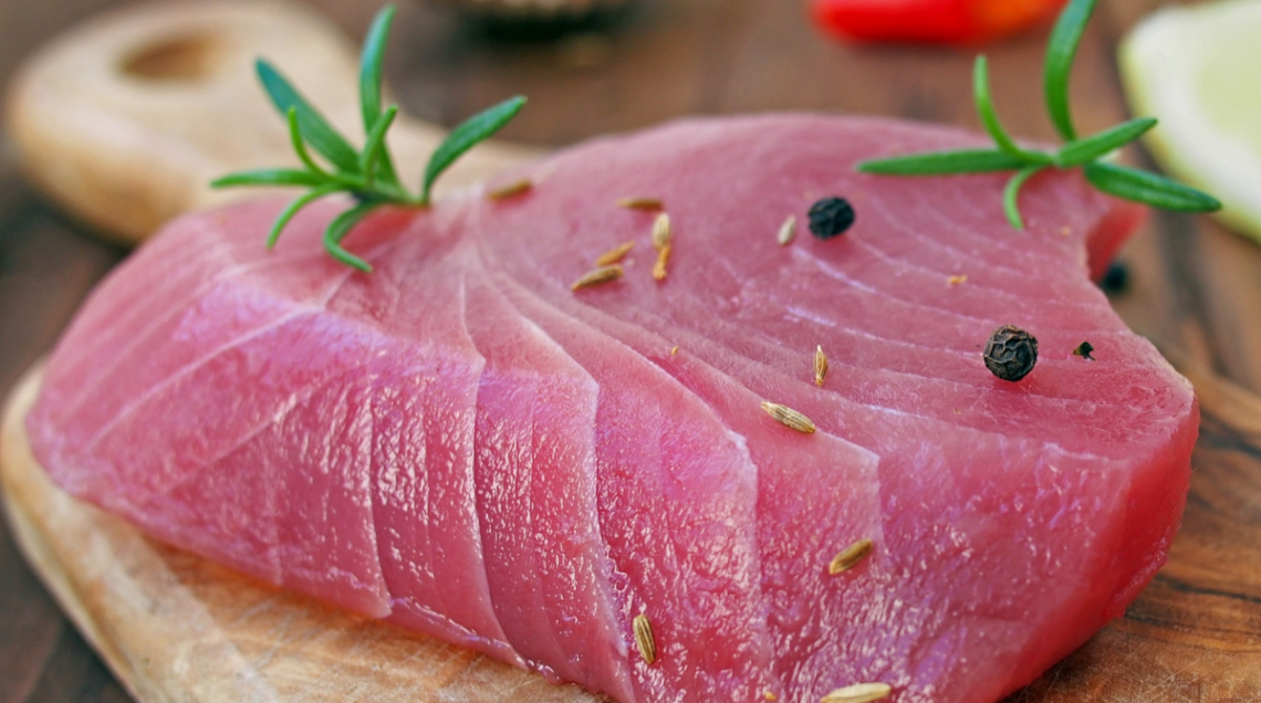 Un filetto fresco e crudo di tonno rosso su un tagliere di legno, guarnito con grani di pepe nero e rametti di rosmarino.