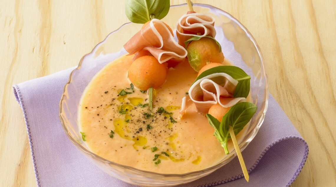 Gazpacho di melone dentro una coppetta trasparente servito con due spedini di melone e prosciutto appoggiati ai bordi